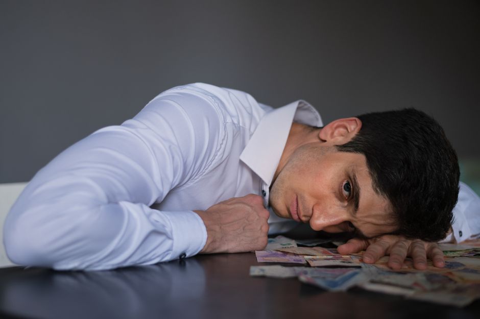 A man in a white dress shirt is lying face down on a dark table, with his head turned to the side, resting on his forearm. His expression appears distressed or exhausted. The table has scattered curre