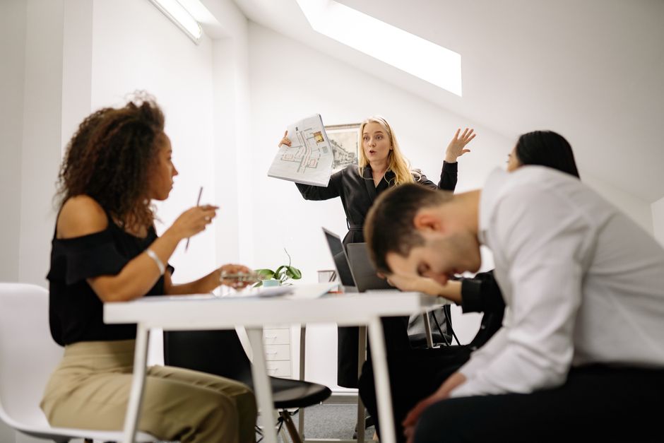 In a modern, well-lit office meeting room, a woman with blonde hair is holding a large newspaper and gesturing animatedly while addressing three colleagues. One woman with curly hair, wearing a black 