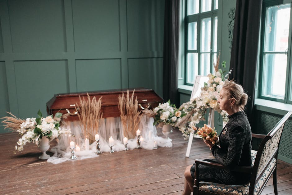 A woman seated indoors on a patterned upholstered chair, holding a bouquet of peach and cream flowers, with an arrangement of white roses, hydrangeas, and greenery on an easel beside her. In the backg