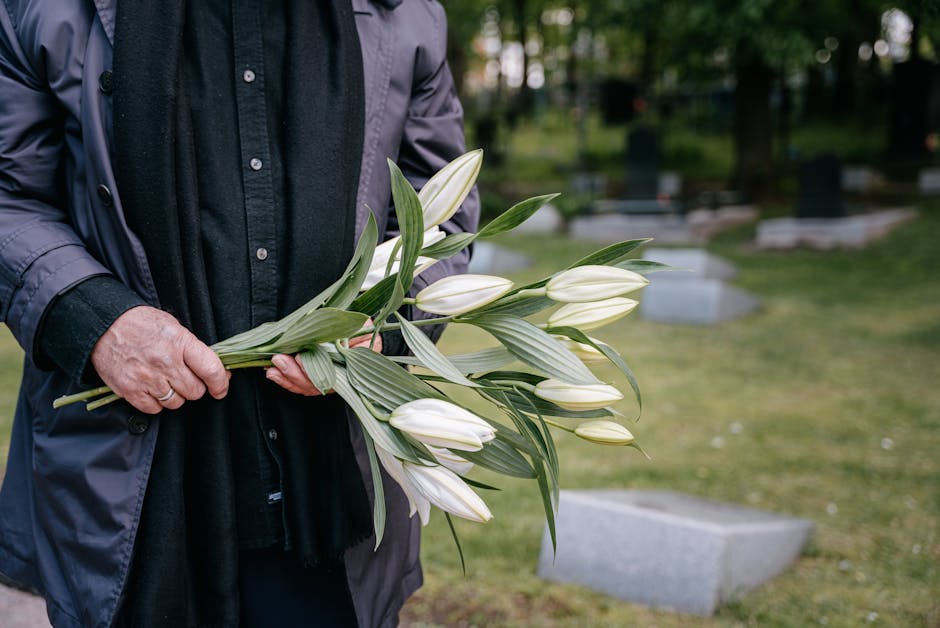 A person wearing a dark jacket and black shirt is holding a bouquet of white lilies with green leaves, their hand gently grasping the stems. The lilies are fresh and closed, with smooth petals and lon